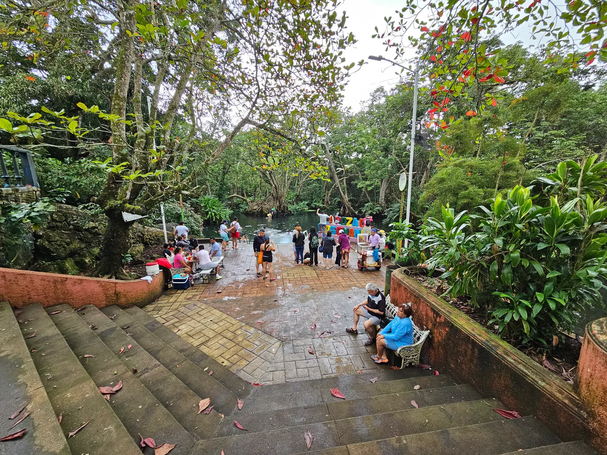 Escaleras que bajan a la Poza de los Enanos entre la vegetación, con visitantes junto al agua de manantial, en Sontecomapan, Veracruz