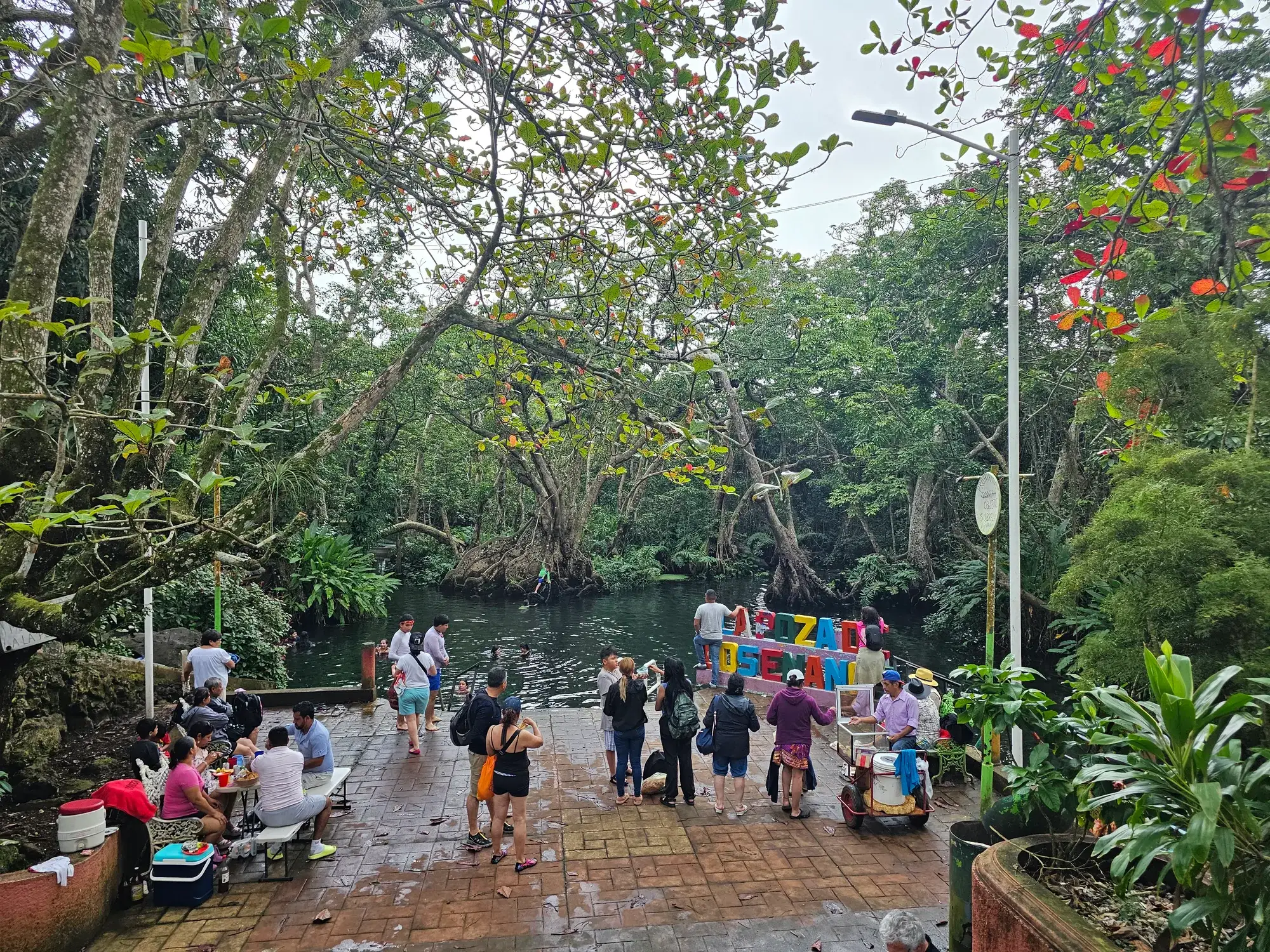 Explanada de la Poza de los Enanos con mesas y puestos de comida junto al agua, rodeada de selva, en Sontecomapan, Veracruz