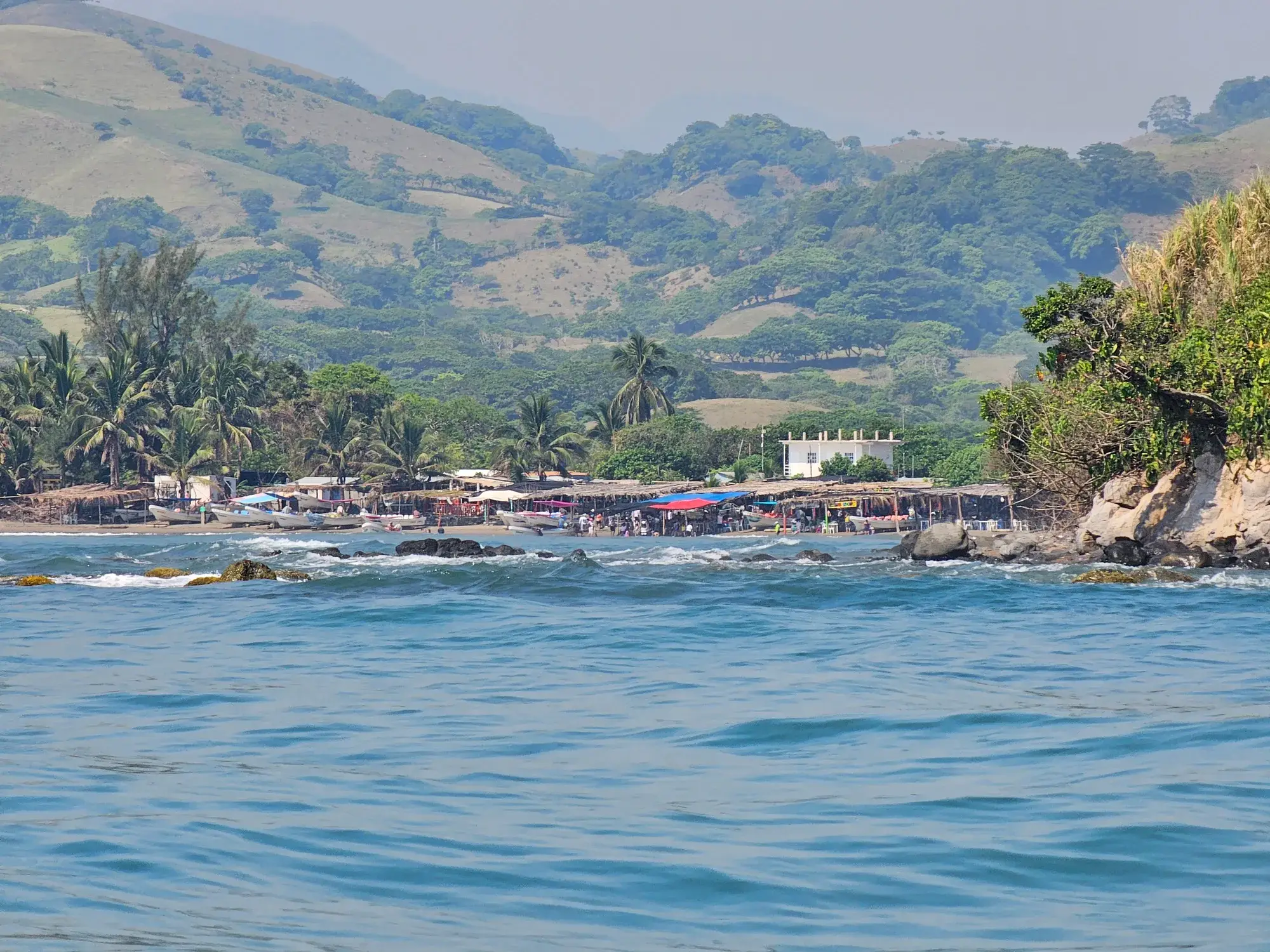 Costa de Los Tuxtlas vista desde la lancha: playa con palapas y palmeras al pie de los cerros verdes, cerca de Roca Partida